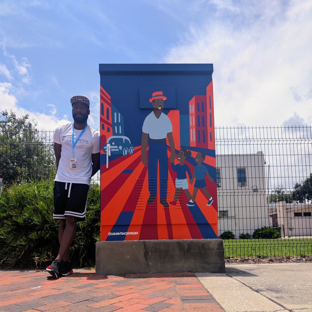 Artist standing beside a vibrant outdoor mural depicting a father walking with two children through a colorful cityscape.